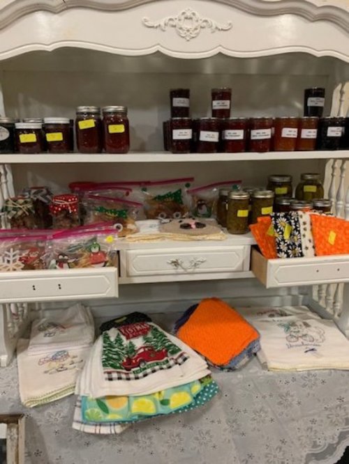 A display of homemade jars, bags of treats, and colorful kitchen towels on a white shelf.