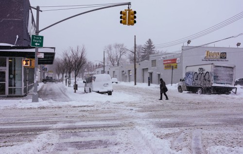A snowy street scene with a traffic light, a person walking, and vehicles partially buried in snow.