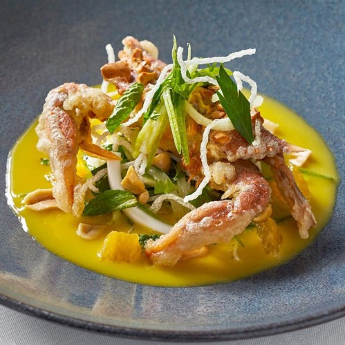 A vibrant dish featuring crispy fried seafood atop a colorful salad with herbs, nuts, and a yellow sauce in a blue bowl.