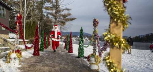 A person dressed as Santa runs along a snowy path decorated with colorful holiday trees and garlands by a lake.