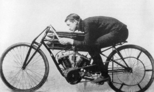 A vintage black and white photo of a man in a racing position on an early motorcycle.