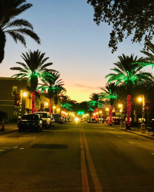 A festive street lined with palm trees adorned with colorful lights, under a sunset sky.