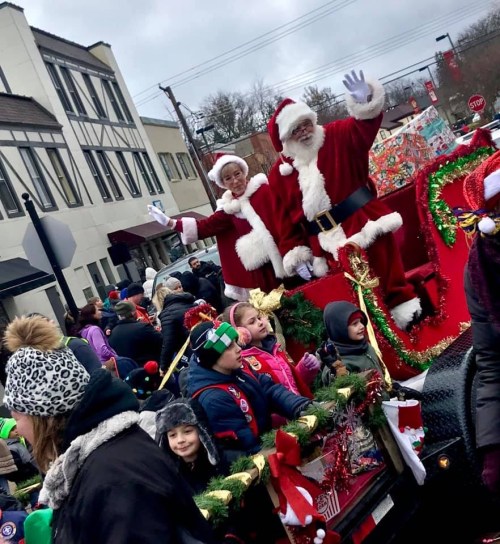 Two Santa Clauses wave from a festive float, surrounded by a crowd of children and holiday decorations.