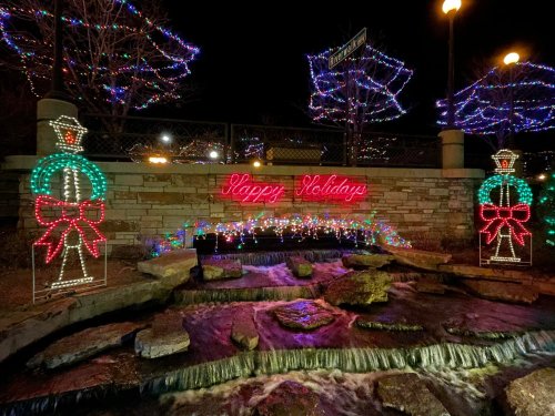 Colorful holiday lights decorate a stone wall and a stream, with a "Happy Holidays" sign in the center.