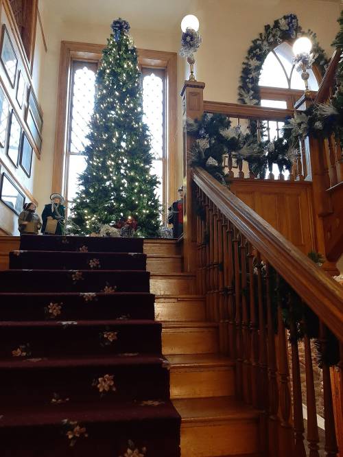 A staircase decorated for the holidays, featuring a lit Christmas tree and garlands, leading to a warmly lit upper area.