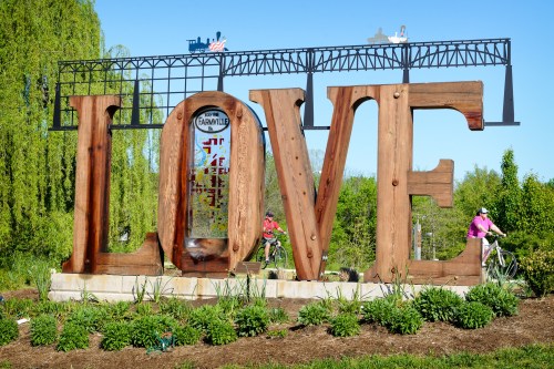 A large wooden sculpture spelling "LOVE" surrounded by greenery, with people on bicycles in the background.