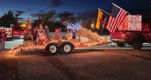 A festive parade float decorated with lights, a Christmas tree, and flags, celebrating the holiday season at dusk.