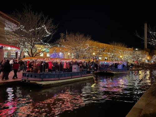A festive night scene with boats on a canal, surrounded by colorful lights and a crowd enjoying the holiday atmosphere.