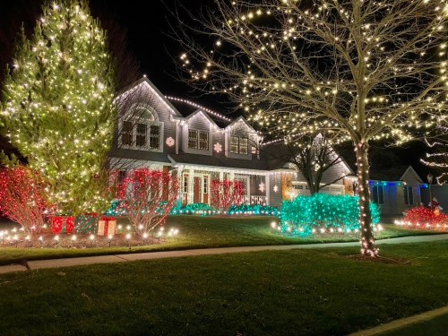 A beautifully decorated house with colorful Christmas lights, surrounded by illuminated trees and festive shrubs at night.