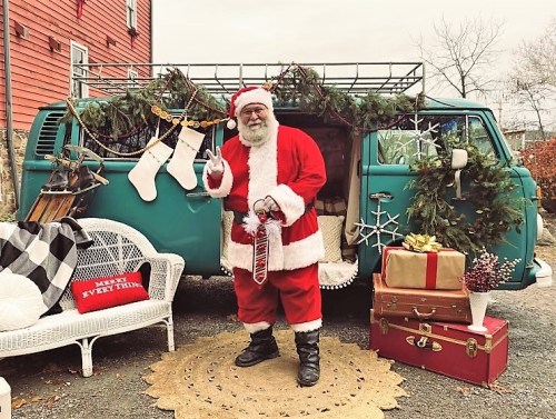 Santa Claus stands beside a decorated vintage van, surrounded by holiday decorations, gifts, and a cozy seating area.