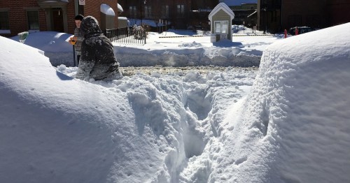 A person shovels snow in a deep snow-covered area, with high snowbanks on either side.