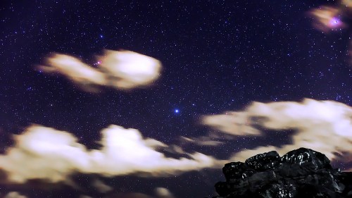 A starry night sky with wispy clouds and a dark rock formation in the foreground.