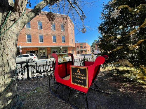 A festive red sleigh with a sign reading "Greetings from Berlin" in a town square, surrounded by holiday decorations.