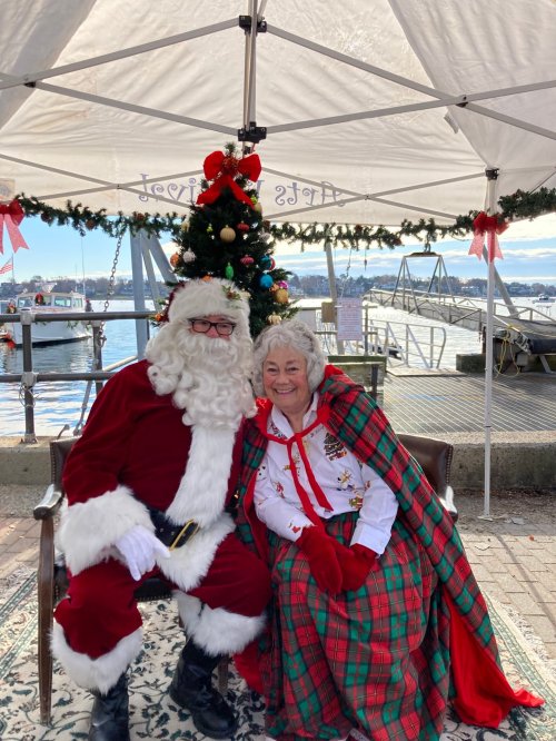 Santa Claus and a woman in festive attire sit together under a decorated tent by the water, smiling joyfully.