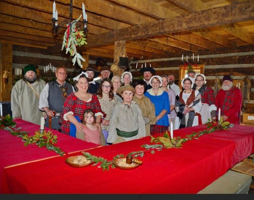 A group of people in historical costumes poses around a festive table in a rustic setting, decorated for a celebration.