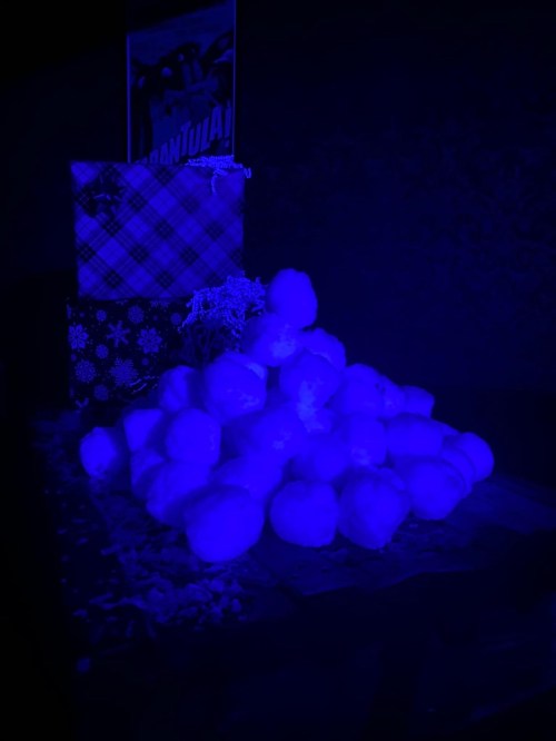 A pile of white snowballs on a dark surface, illuminated in blue light, with wrapped gifts in the background.