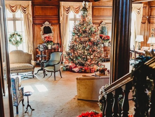 A cozy living room decorated for the holidays, featuring a large Christmas tree surrounded by gifts and elegant furnishings.