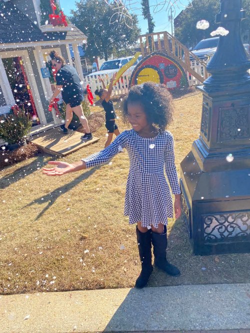 A girl in a checkered dress reaches out as snowflakes fall around her, with festive decorations in the background.