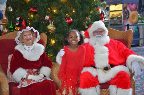 A young girl in a red dress smiles with Santa and Mrs. Claus in front of a decorated Christmas tree.