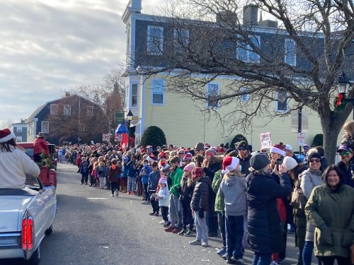 A large crowd of people in winter clothing, many wearing Santa hats, lined up along a street for a parade.