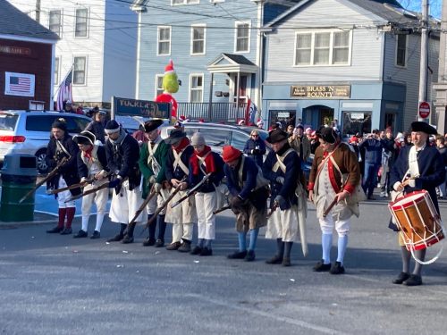 A group of historical reenactors in colonial attire, lined up with muskets, during a festive outdoor event.