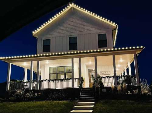 A beautifully lit two-story house at night, adorned with string lights along the roof and porch.