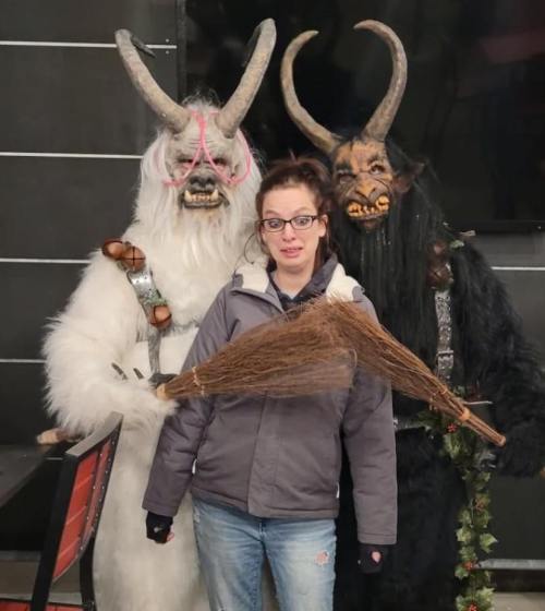 A woman stands between two costumed figures with horns, one in white fur and the other in black, holding a broom.