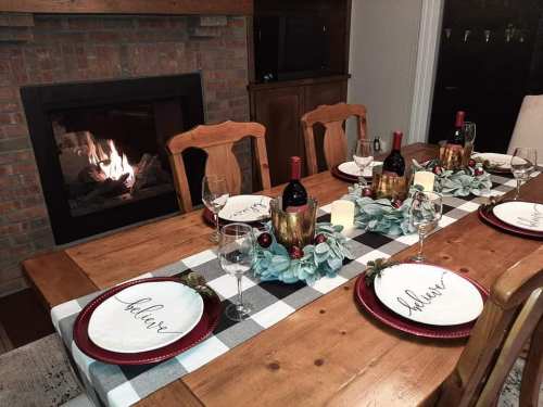 A cozy dining table set for a festive meal, featuring wine bottles, elegant plates, and a warm fireplace in the background.