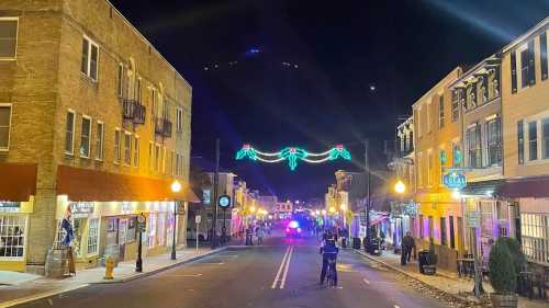 A festive street at night, adorned with holiday lights and decorations, showcasing shops and a police vehicle in the distance.