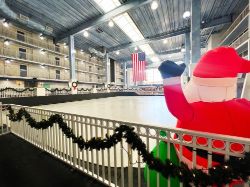 A large inflatable Santa waves beside an empty ice rink, decorated with garlands and an American flag in the background.