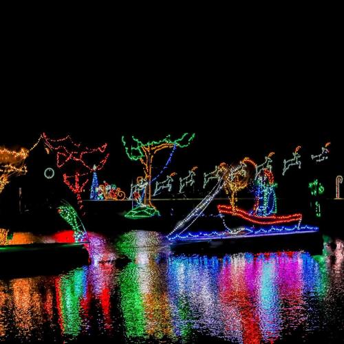 Colorful holiday lights illuminate trees and figures along a waterway, reflecting beautifully on the surface at night.