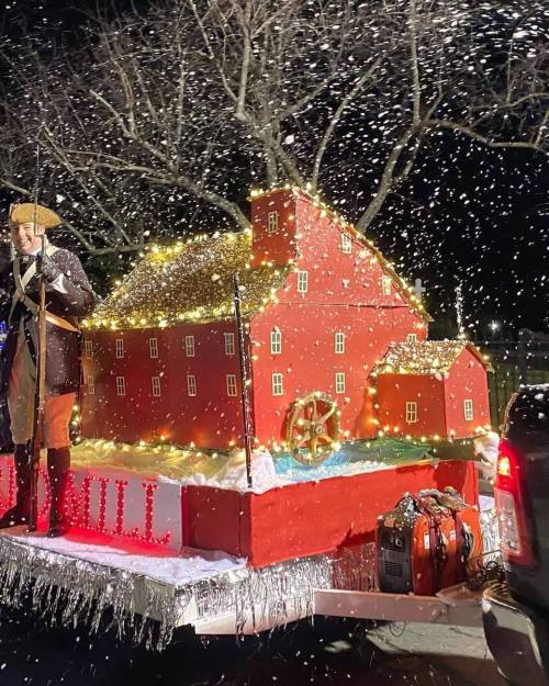 A festive parade float featuring a large red barn adorned with lights, snow, and a person in historical costume.