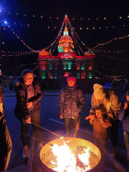 A group of children and adults gather around a fire pit, with a brightly lit building and festive lights in the background.