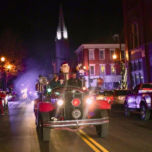 A vintage fire truck drives down a festive street, with Santa waving from the passenger seat and holiday lights in the background.