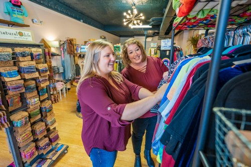 Two women shopping in a boutique, smiling as they browse colorful clothing on a rack.