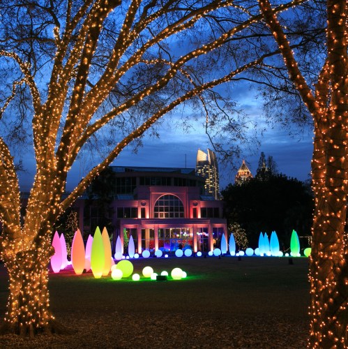 Colorful lights illuminate trees and decorative shapes in a park, with a city skyline visible in the background at night.