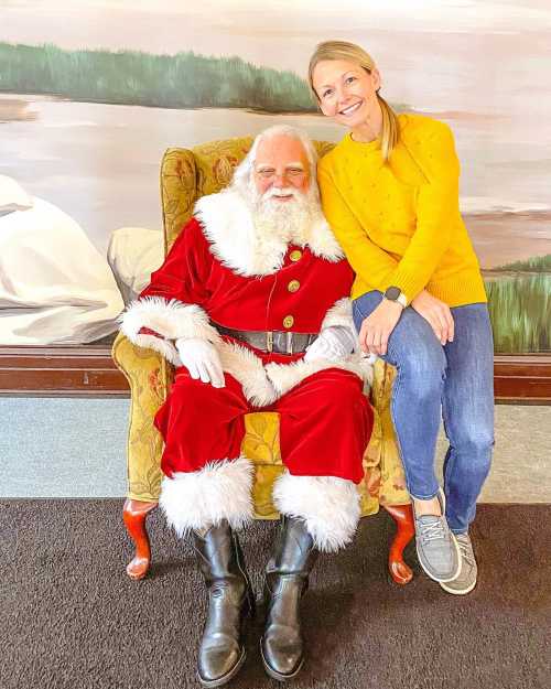 A smiling woman in a yellow sweater sits next to Santa Claus in a festive setting.