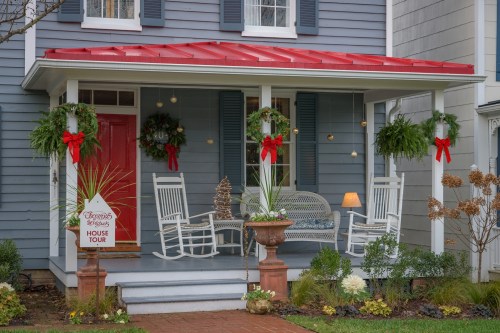 A charming house with a red roof, decorated for the holidays, featuring wreaths, rocking chairs, and a sign for a house tour.