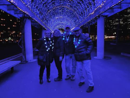 Four people stand under a blue-lit archway, wearing glowing necklaces, with city lights in the background.