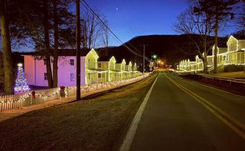 A festive street lined with illuminated houses and a decorated Christmas tree, set against a twilight sky.