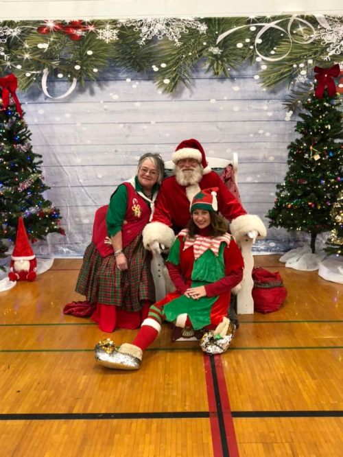 A woman in festive attire and an elf pose with Santa Claus in front of decorated Christmas trees and a snowy backdrop.