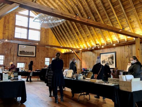 People browse tables filled with books in a spacious, wooden room with high ceilings and large windows.