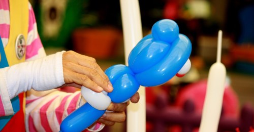 A person’s hand holds a blue balloon animal, with white accents, in a colorful setting filled with other balloons.