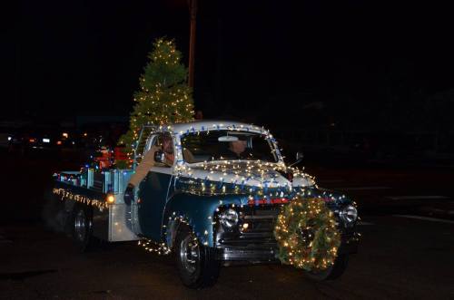 A vintage truck decorated with Christmas lights and a tree, driving at night with festive gifts in the back.