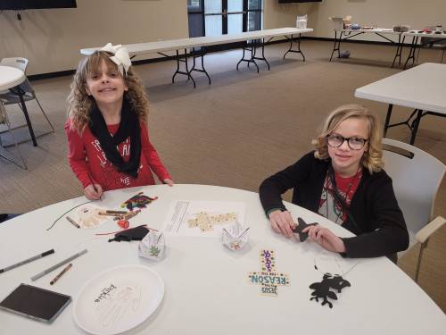 Two children sit at a table, smiling while working on craft projects with various art supplies around them.