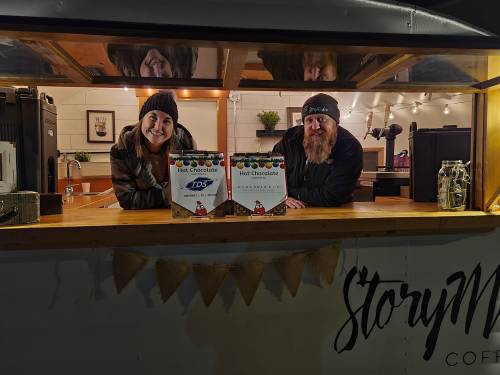 Two people smile from a food truck window, showcasing hot chocolate menus in a cozy, warmly lit setting.