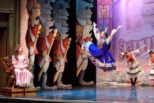 A dancer in a blue dress leaps gracefully on stage, while another girl sits nearby, surrounded by festive decorations.