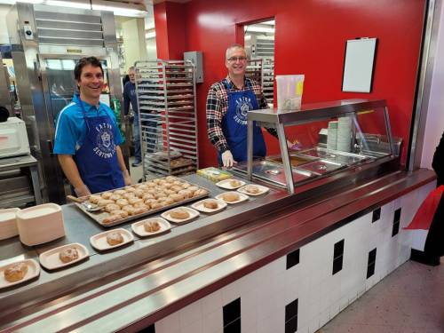 Two smiling men in a cafeteria serving pastries from a counter, with trays of food and a red wall in the background.