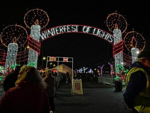 A festive archway with "Winterfest of Lights" illuminated, surrounded by colorful lights and holiday decorations at night.