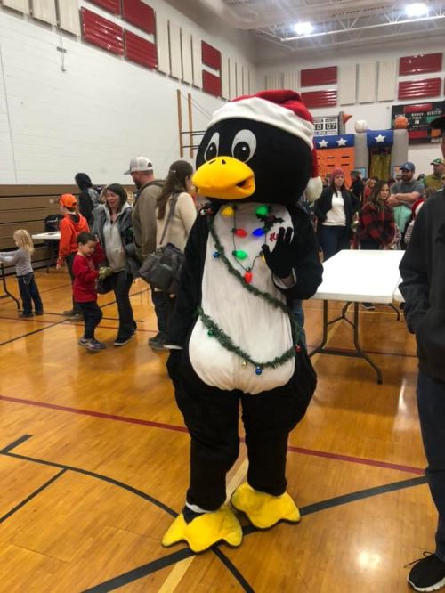 A person in a penguin costume wearing a Santa hat and festive decorations stands in a gym filled with people.
